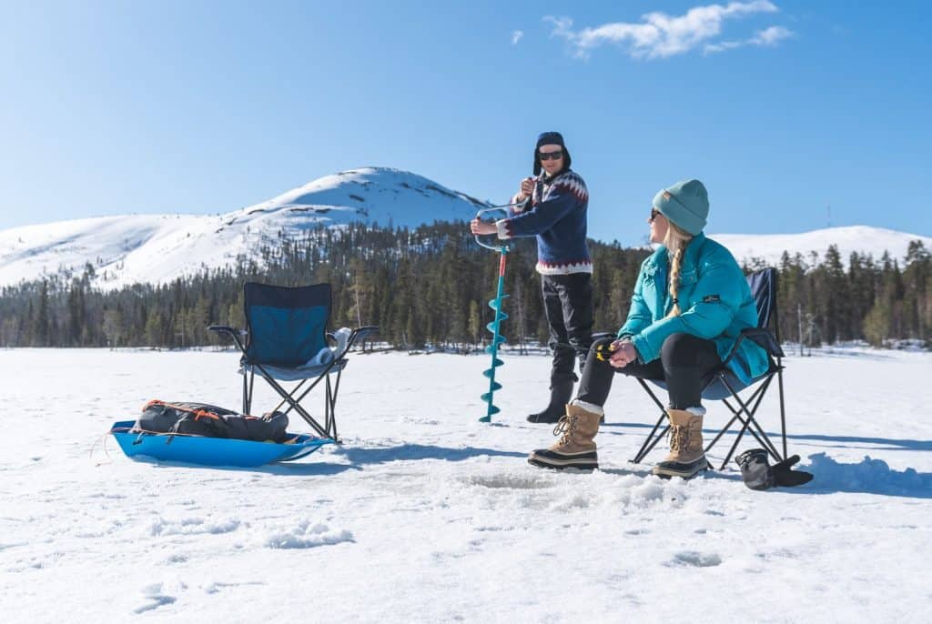 A person using a portable ice auger on snow with two chairs and a mountain in the background.