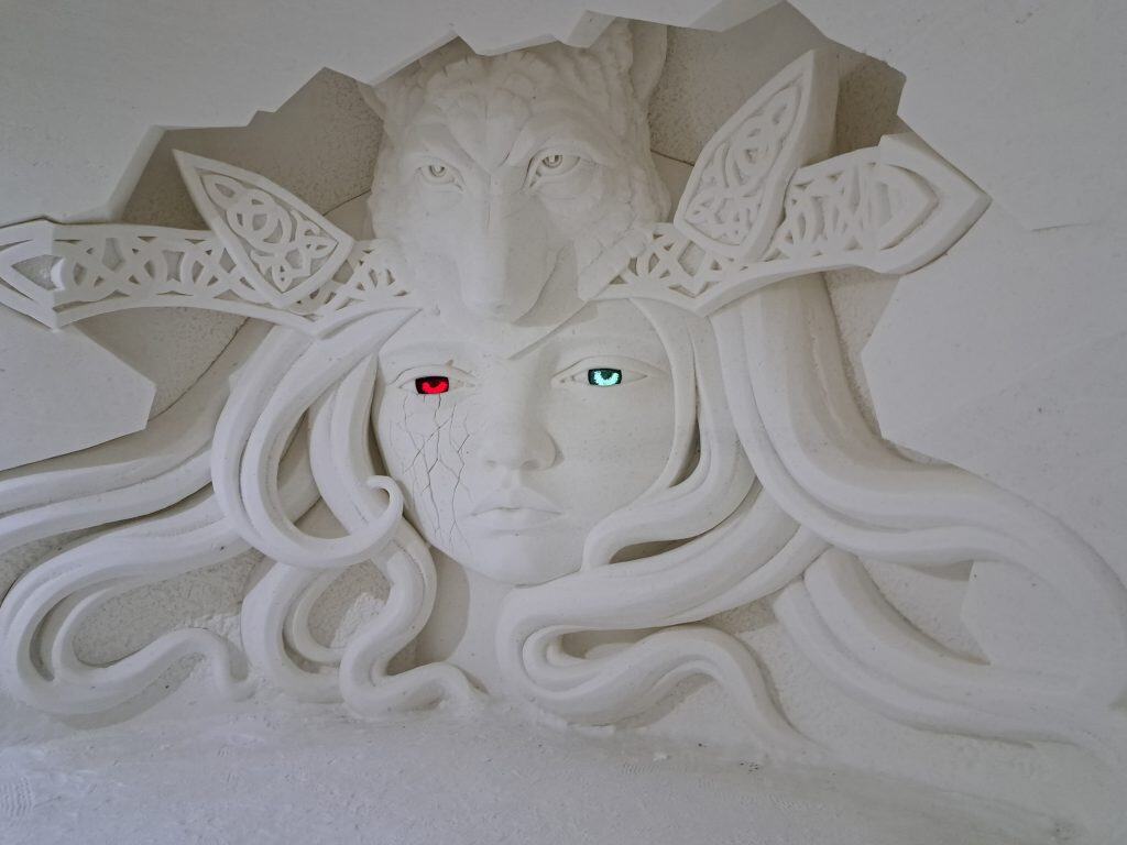A snow sculpture of a woman with flowing hair, a wolf head above, and colourful eyes.