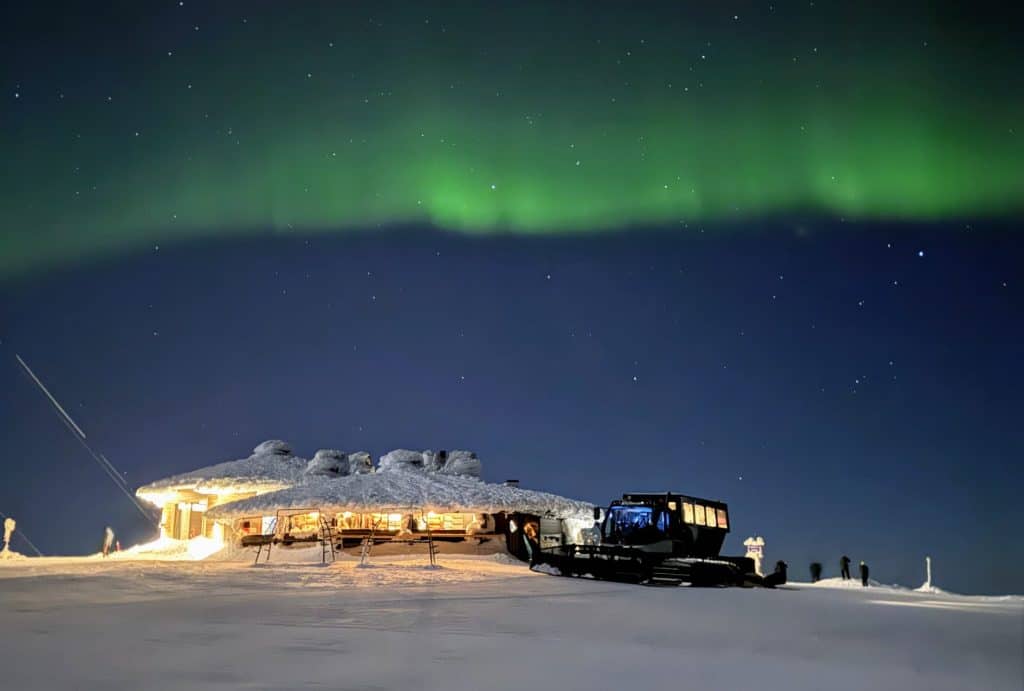 Aurora Borealis over Ylläsjärvi, Finnish Lapland with snow-covered buildings and visitors.
