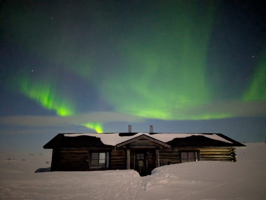 Northern Lights over a snow-covered wooden cabin in Finnish Lapland.