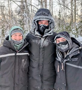 Three people in winter gear smiling in snowy forest in Lapland.